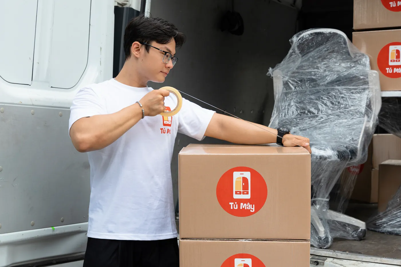 A crew member taping a Tủ Mây box shut, a shrink-wrapped chair already loaded behind