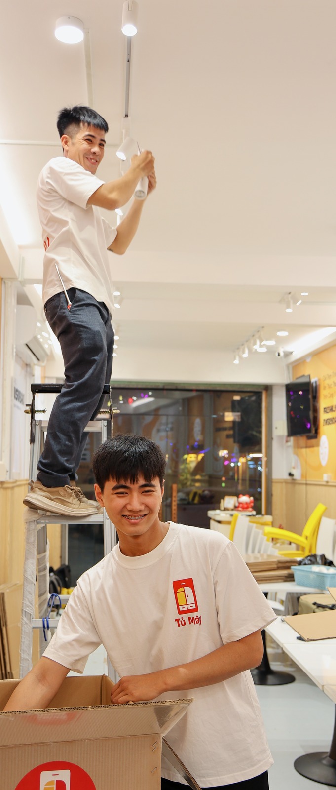 One crew member on a ladder taking down a ceiling light, another holding a box below
