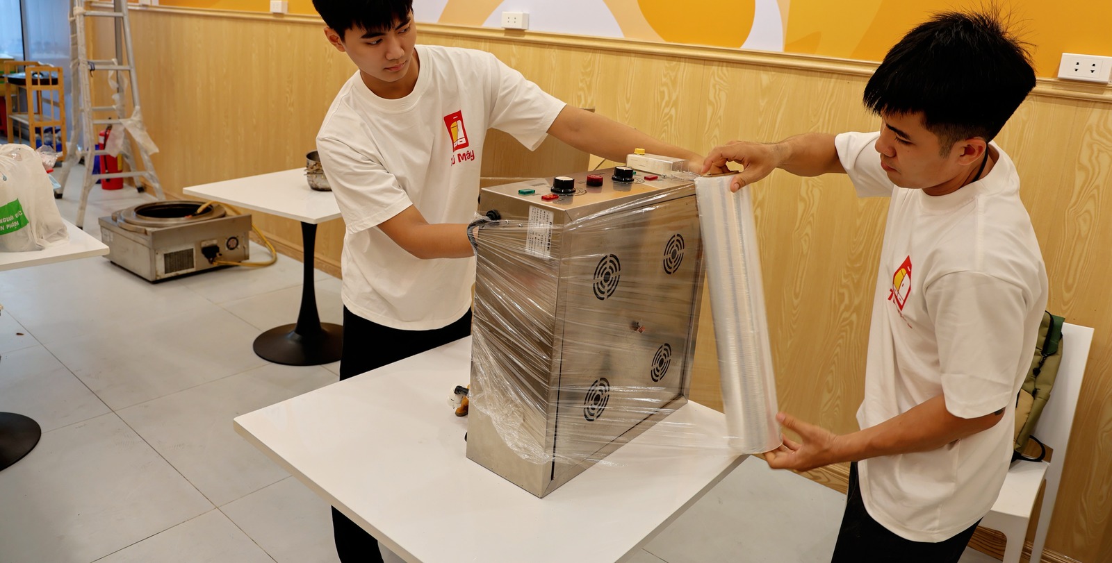 Two crew members wrapping stretch film around a stainless steamer cabinet