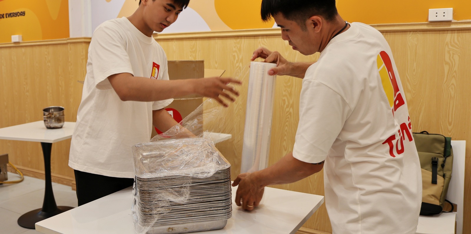 Two crew members wrapping stretch film around a stack of flat trays