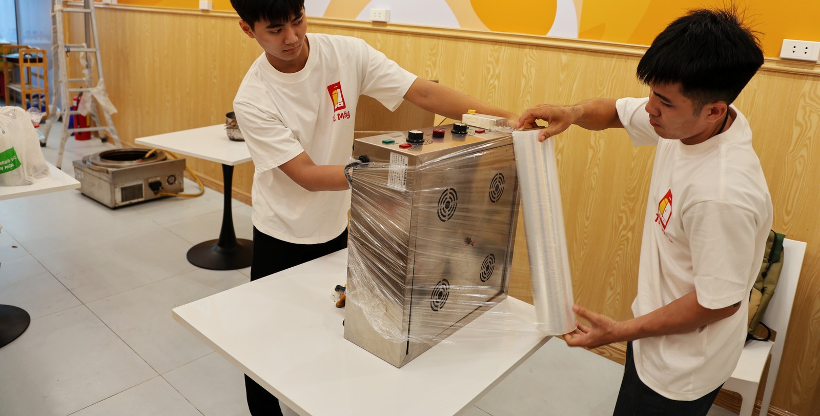 Two crew members wrapping stretch film around a small stainless machine